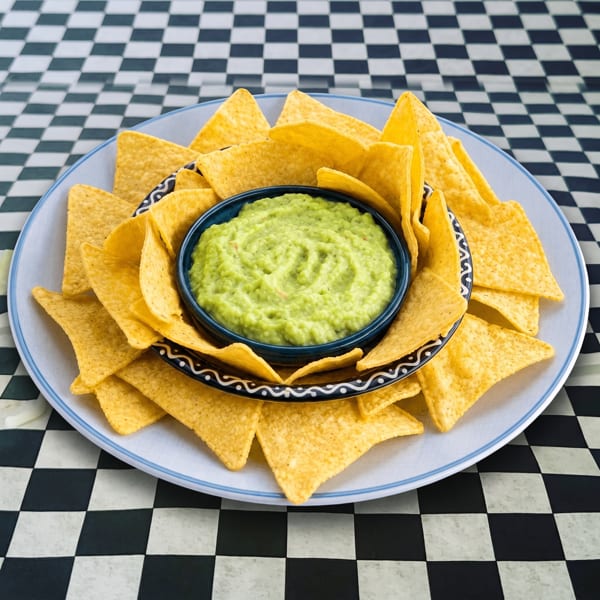 A blue bowl of guacamole surrounded by tortilla chips on a white plate, on a black and white checkered tablecloth.