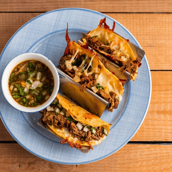 Three cheesy birria tacos with a side of consommé on a blue plate.