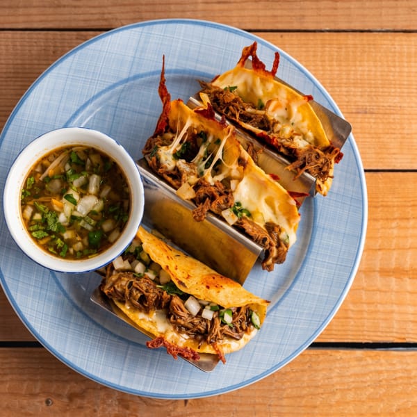 Three cheesy birria tacos with a side of consommé on a blue plate, on a wooden table.