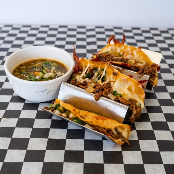 Three cheesy birria tacos with a side of consommé on a black and white checkered paper.